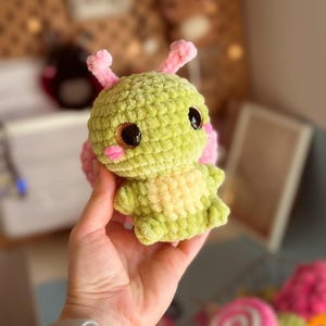 May include: A handmade, plush crochet creature in shades of green and pink. The toy has large, dark eyes, rosy cheeks, and small antennae. The body is a textured, fuzzy material, and the overall design is whimsical and cute.