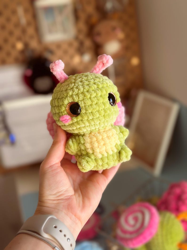 May include: A handmade, plush crochet creature in shades of green and pink. The toy has large, dark eyes, rosy cheeks, and small antennae. The body is a textured, fuzzy material, and the overall design is whimsical and cute.