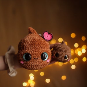 Puede incluir: Dos juguetes de peluche de crochet con forma de caca con caras lindas. El más grande es marrón con un corazón rosa en la parte superior y ojos azules. El más pequeño es marrón con ojos negros.