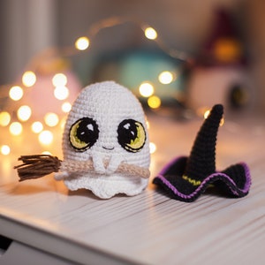 Pu&ograve; includere: Un fantasma bianco all'uncinetto che tiene una scopa marrone con un viso giallo e occhi grandi. Un cappello da strega nero all'uncinetto con un bordo viola si trova accanto al fantasma.