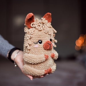 May include: A handmade plush llama toy in shades of tan and brown. The llama has a fluffy body, small ears, and a smiling face with pink cheeks. The toy is held in a person's hand.