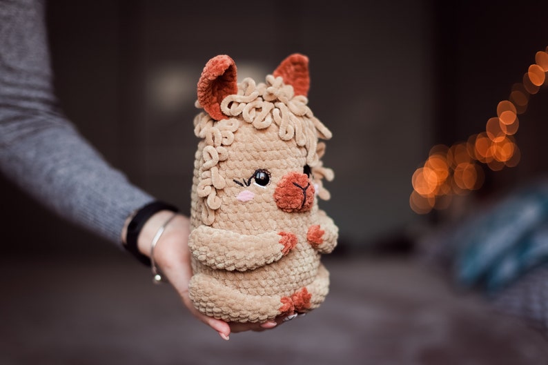 May include: A handmade plush llama toy in shades of tan and brown. The llama has a fluffy body, small ears, and a smiling face with pink cheeks. The toy is held in a person's hand.