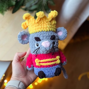 May include: A crocheted mouse toy, featuring a yellow crown and a red jacket with gold detailing. The mouse has grey fur, pink ears, and a pink nose. The toy is held in a person's hand.