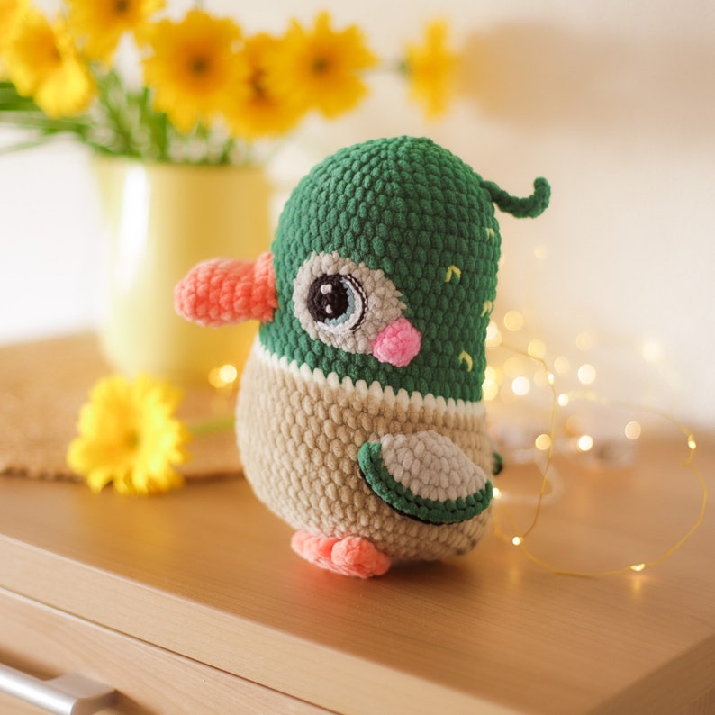 May include: A crocheted duck plush toy with green and beige yarn. The duck has a pink beak and large blue eyes. The toy is sitting on a wooden surface.