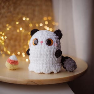May include: A white crocheted ghost with black ears and a gray tail. The ghost has large, sparkly eyes and a pink nose. The ghost is sitting on a wooden table with a candy corn crocheted toy in the foreground.
