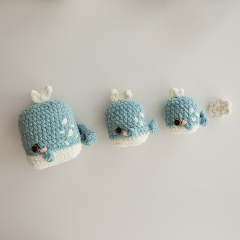 May include: Three crocheted whale plushies in different sizes, all blue with white accents. The largest whale is on the left, the smallest on the right.