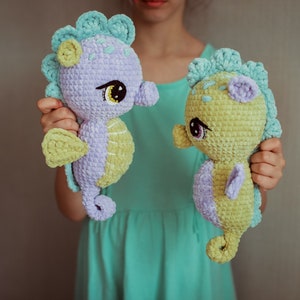 May include: Two crocheted seahorse plushies, one purple and one yellow, with green accents. The seahorses have large, black eyes and are held by a person.