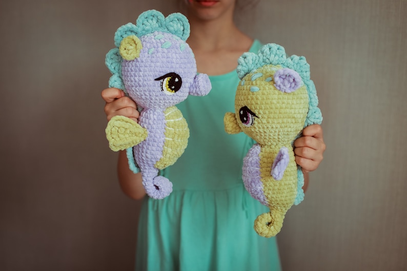 May include: Two crocheted seahorse plushies, one purple and one yellow, with green accents. The seahorses have large, black eyes and are held by a person.