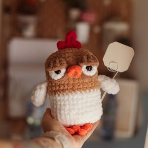 May include: A crocheted chicken toy with brown and white fur, red comb, and orange feet. The chicken is holding a blank white tag.