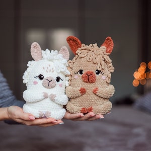May include: Two handmade crochet llama dolls. One is white, the other is tan. Both have pink accents on their ears and cheeks, with black eyes and a small, embroidered mouth. The dolls are held in a person's hands.