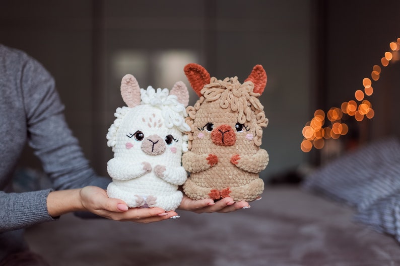 May include: Two handmade crochet llama dolls. One is white, the other is tan. Both have pink accents on their ears and cheeks, with black eyes and a small, embroidered mouth. The dolls are held in a person's hands.