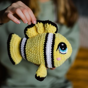 May include: A crocheted yellow and black fish toy with white stripes and large blue eyes. The fish is being held by a person's hand.