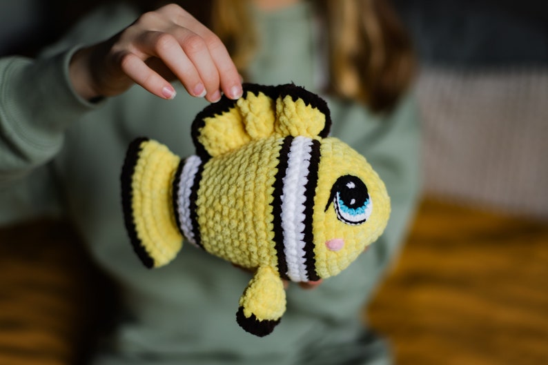 May include: A crocheted yellow and black fish toy with white stripes and large blue eyes. The fish is being held by a person's hand.