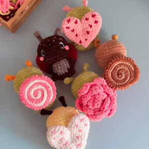 May include: A collection of handmade crochet items, including a ladybug, hearts, and floral designs. The items are in various colors, such as pink, yellow, brown, and green. A wooden crate filled with yarn is in the background.