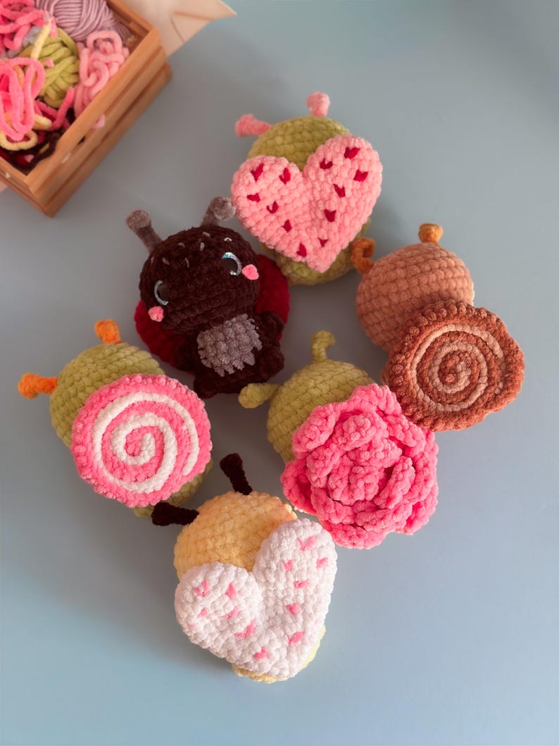 May include: A collection of handmade crochet items, including a ladybug, hearts, and floral designs. The items are in various colors, such as pink, yellow, brown, and green. A wooden crate filled with yarn is in the background.