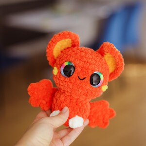 Puede incluir: Un murci&eacute;lago naranja de crochet con orejas amarillas y grandes ojos arco&iacute;ris. El murci&eacute;lago sostiene un coraz&oacute;n blanco de crochet en sus patas.