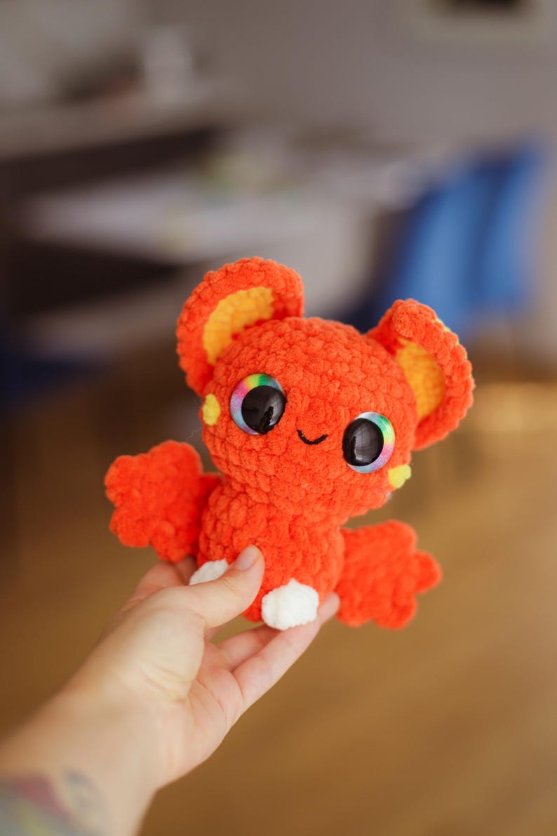 Puede incluir: Un murci&eacute;lago naranja de crochet con orejas amarillas y grandes ojos arco&iacute;ris. El murci&eacute;lago sostiene un coraz&oacute;n blanco de crochet en sus patas.