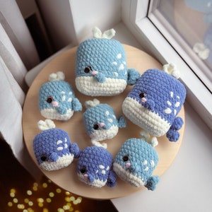 以下が含まれることがあります： 青と白の色合いで作られた、7匹の編みぐるみクジラの人形セット。クジラは木製の台の上に並べられています。