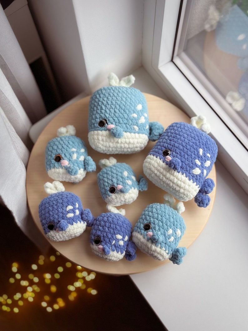 May include: A set of seven crocheted whale plushies in shades of blue and white. The whales are arranged on a wooden surface.