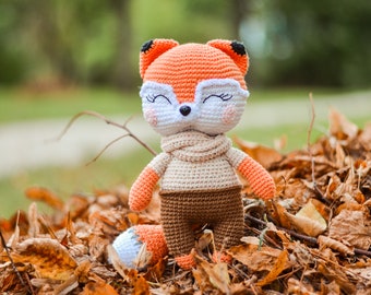 Puede incluir: Una muñeca de zorro de ganchillo con hilo naranja, blanco y marrón. El zorro tiene una cara blanca con los ojos cerrados, una bufanda beige y pantalones marrones. La muñeca está sentada sobre un lecho de hojas de otoño.