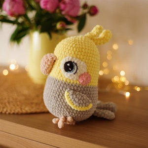 May include: A crocheted stuffed bird toy with yellow and gray yarn. The bird has a small beak, large eyes, and a small pink cheek.