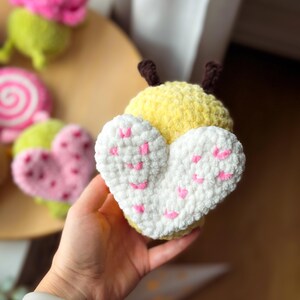 May include: A handmade yellow and white crochet butterfly with pink heart accents. The butterfly is held in a hand, with other crochet items in the background, including a pink flower, a green and pink heart, and a pink and white swirl.