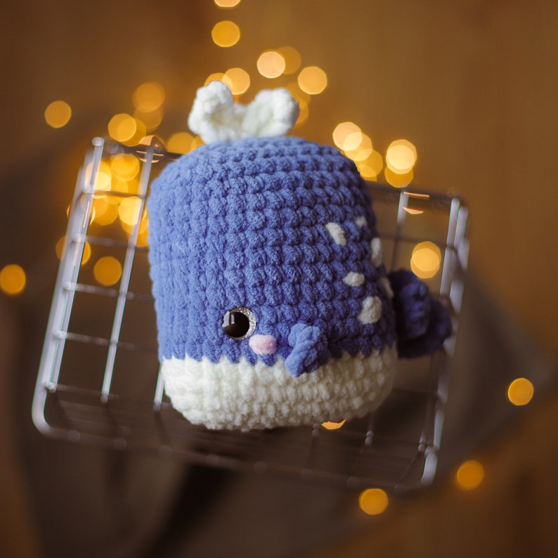 May include: A crocheted whale plush toy in blue and white with a white spout and a black eye. The whale is sitting on a metal rack with a blurred background of lights.