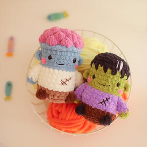 May include: Two small, plush, crocheted figures sit in a wire basket. One figure has a light blue body, a pink brain-like top, and a white shirt with a brown base. The other figure is green with black hair, a purple shirt, and a brown base.