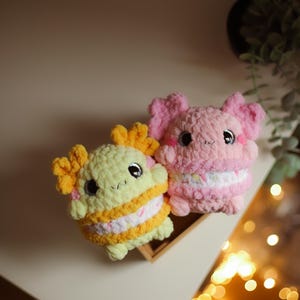 Puede incluir: Dos peluches de crochet con forma de ajolote. Uno es amarillo con detalles naranjas y el otro es rosa. Ambos tienen ojos negros grandes y una franja blanca alrededor de la mitad.