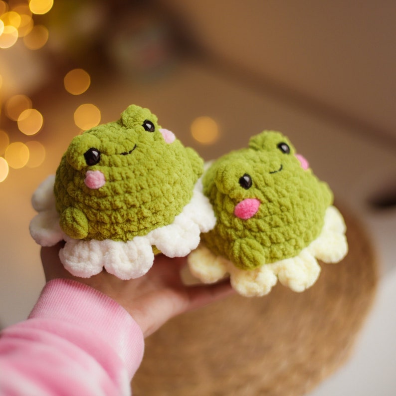 May include: Two crocheted green frog plushies with white and yellow accents. The frogs have pink cheeks and smiling faces.
