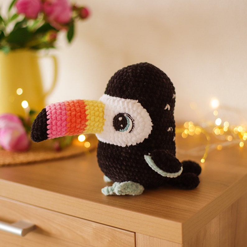May include: A crocheted toucan plush toy with a black body, white face, and a multi-colored beak. The toucan is sitting on a wooden surface.