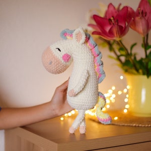 Può includere: Un giocattolo di unicorno all'uncinetto bianco con accenti rosa, blu e gialli. L'unicorno ha una criniera e una coda soffici e si trova su una superficie di legno.