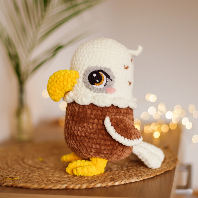 May include: A crocheted eagle plush toy with brown and white fur, yellow beak, and large black eyes. The toy is sitting on a woven brown surface.