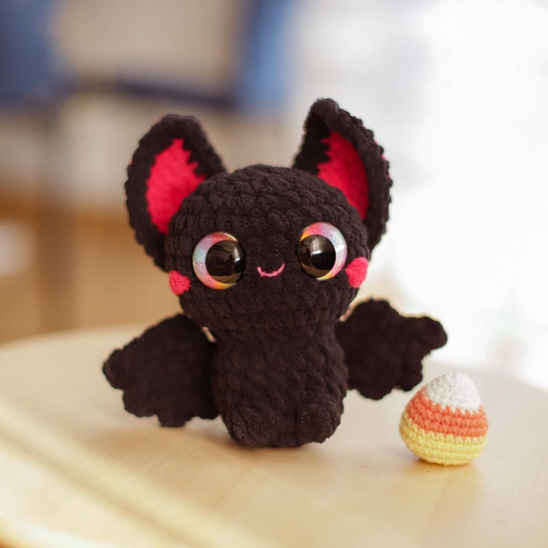 Puede incluir: Un murci&eacute;lago de crochet negro con orejas rosas y ojos iridiscentes. El murci&eacute;lago sostiene un caramelo de ma&iacute;z de crochet.