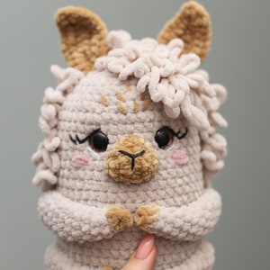 May include: A handmade, light beige crochet llama plush toy with brown eyes, eyelashes, and pink cheeks. The llama has fluffy, light beige yarn around its head and ears. The llama's nose and inner ears are a darker brown. The llama's hands are clasped together.