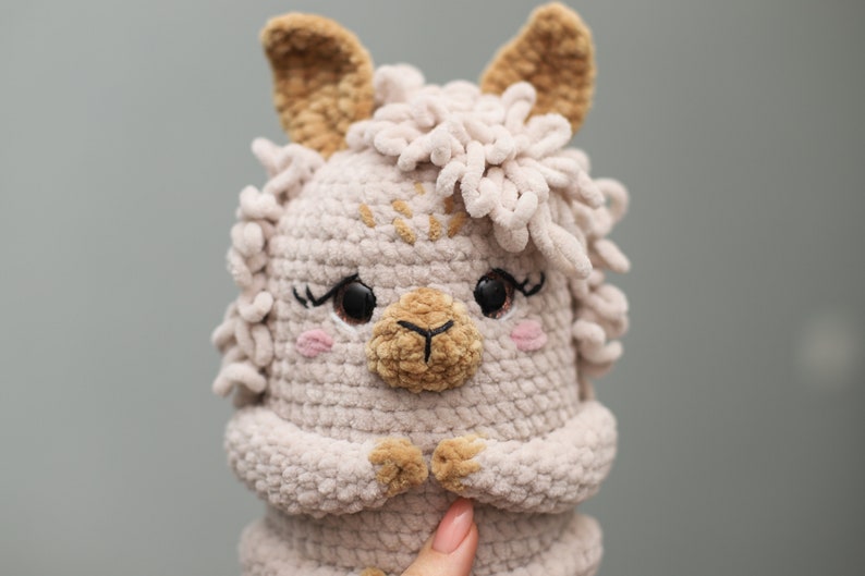 May include: A handmade, light beige crochet llama plush toy with brown eyes, eyelashes, and pink cheeks. The llama has fluffy, light beige yarn around its head and ears. The llama's nose and inner ears are a darker brown. The llama's hands are clasped together.