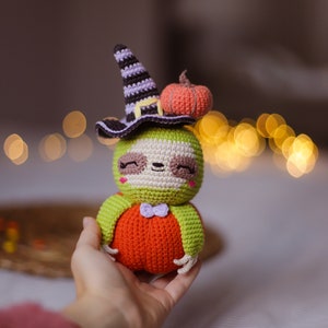 May include: A crocheted sloth wearing a witch hat and holding a small orange pumpkin. The sloth is green and white with a purple bow tie.