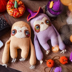 Op de afbeelding: Twee gehaakte luiaard knuffels, een bruin en een paars, met grote ogen en glimlachende gezichten. De bruine luiaard heeft een beige buik en de paarse luiaard draagt een heksenhoed. De knuffels zijn omringd door garenbollen en andere gehaakte items.