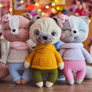 Puede incluir: Tres animales de peluche de crochet: un zorro rosa con la cara blanca, un pug beige con un suéter amarillo y un gato gris con un suéter morado. Los animales llevan todos bufandas y tienen ojos bordados.