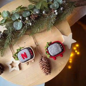 以下が含まれることがあります： 木製のクリスマスオーナメントディスプレイ。画面に文字「N」が入ったかぎ針編みのテレビオーナメント2つ、松ぼっくり、星型のオーナメントが特徴です。ディスプレイは緑と銀色のベリーで飾られています。