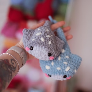 Op de afbeelding: Twee handgemaakte gehaakte roggen, één grijs en één blauw, elk met witte stippen en roze wangen. De roggen hebben zwarte ogen en worden vastgehouden voor een wazige achtergrond van andere kleurrijke haakartikelen.