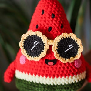 Puede incluir: Un juguete de peluche de sand&iacute;a rojo tejido a crochet con gafas de sol amarillas y negras. La sand&iacute;a tiene una c&aacute;scara verde y una pulpa blanca.
