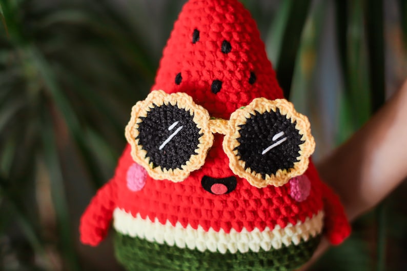 Puede incluir: Un juguete de peluche de sand&iacute;a rojo tejido a crochet con gafas de sol amarillas y negras. La sand&iacute;a tiene una c&aacute;scara verde y una pulpa blanca.