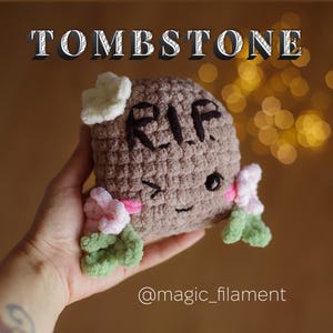 Puede incluir: Un juguete de peluche con forma de lápida crocheteado con una flor blanca en la parte superior, una flor rosa a cada lado y la palabra "RIP" escrita en la parte delantera. El juguete tiene una cara sonriente con ojos negros y una nariz rosa.