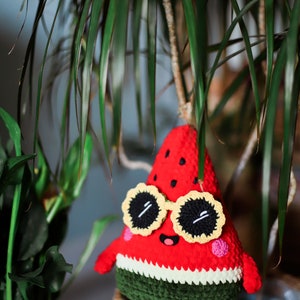 Puede incluir: Un juguete de peluche de sand&iacute;a tejido a crochet que lleva gafas de sol amarillas con lentes negros. La sand&iacute;a es roja con semillas negras y piel verde. El juguete est&aacute; sentado en una cesta tejida.