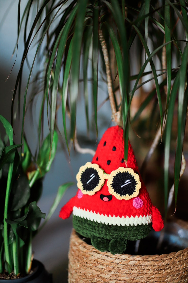 Puede incluir: Un juguete de peluche de sand&iacute;a tejido a crochet que lleva gafas de sol amarillas con lentes negros. La sand&iacute;a es roja con semillas negras y piel verde. El juguete est&aacute; sentado en una cesta tejida.
