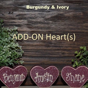 May include: Three burgundy heart-shaped ornaments with ivory glitter and names "Benjamin", "Austin", and "Shane" written in ivory. The hearts are on a dark wood background with greenery on either side. The text "ADD-ON Heart(s)" is above the hearts and "Burgundy & Ivory" is at the top of the image.