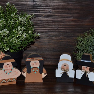 May include: Four wooden cutout figures depicting two individuals in tan and brown attire with headbands and two individuals in black and white pilgrim-style clothing with hats, all featuring painted facial details and small twine accents.