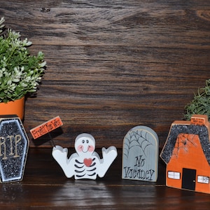 May include: A set of four Halloween-themed wooden decorations including a black coffin with glitter and the text RIP, a white skeleton figure holding a sign that reads Fright This Way, a gray tombstone with spiderwebs and No Vacancy, and an orange house.