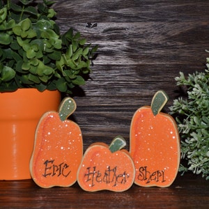 May include: Three orange wooden pumpkin decorations with green stems and glitter accents. Each pumpkin has a name written on it: Eric, Heather, and Sheri. A potted plant and greenery are in the background.